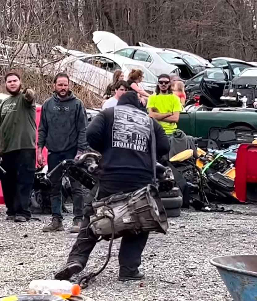 People looking at someone carry a transmission on his back at the junkyard